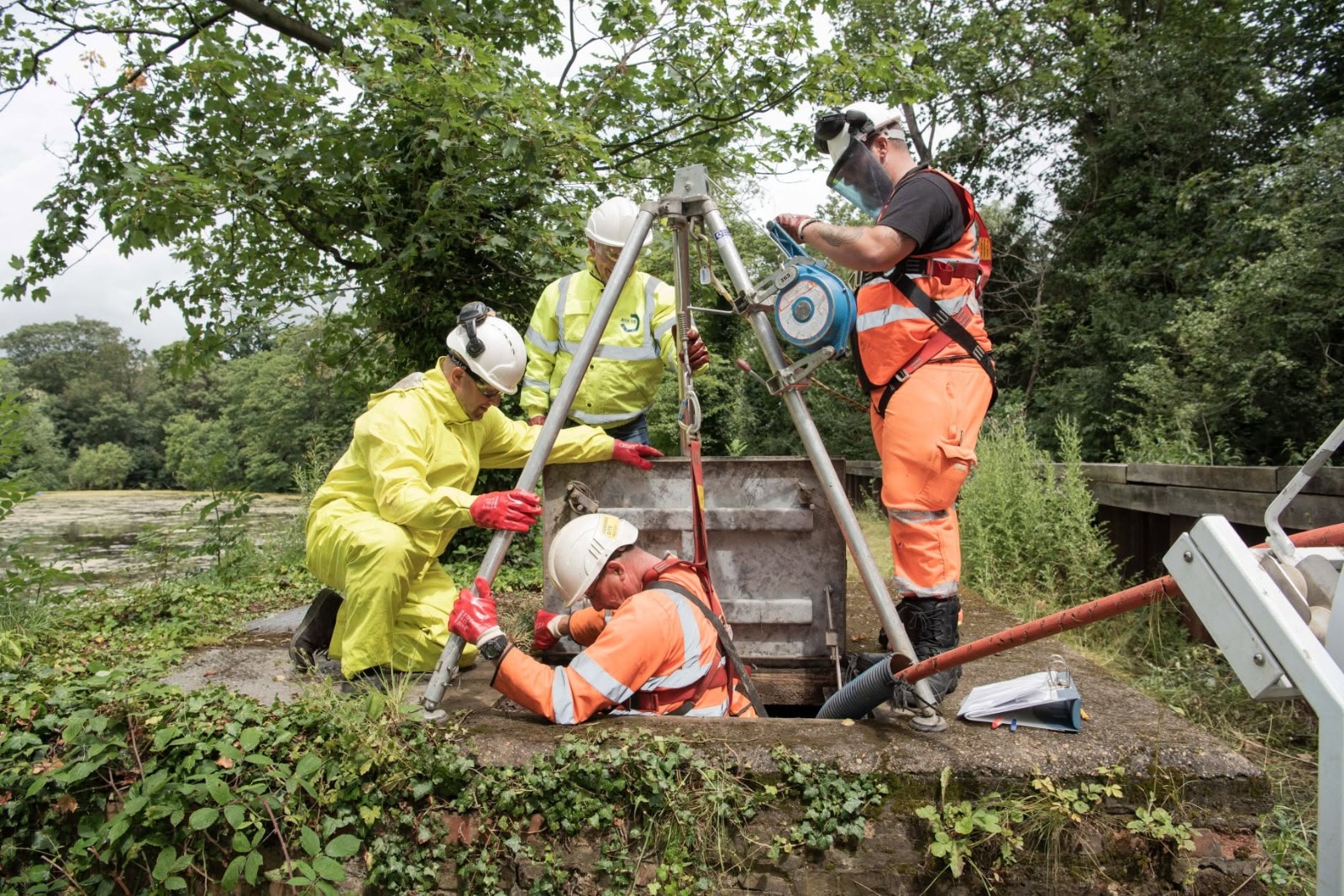 Confined-space drainage team replacing a manhole cover using a tripod hoist and full PPE